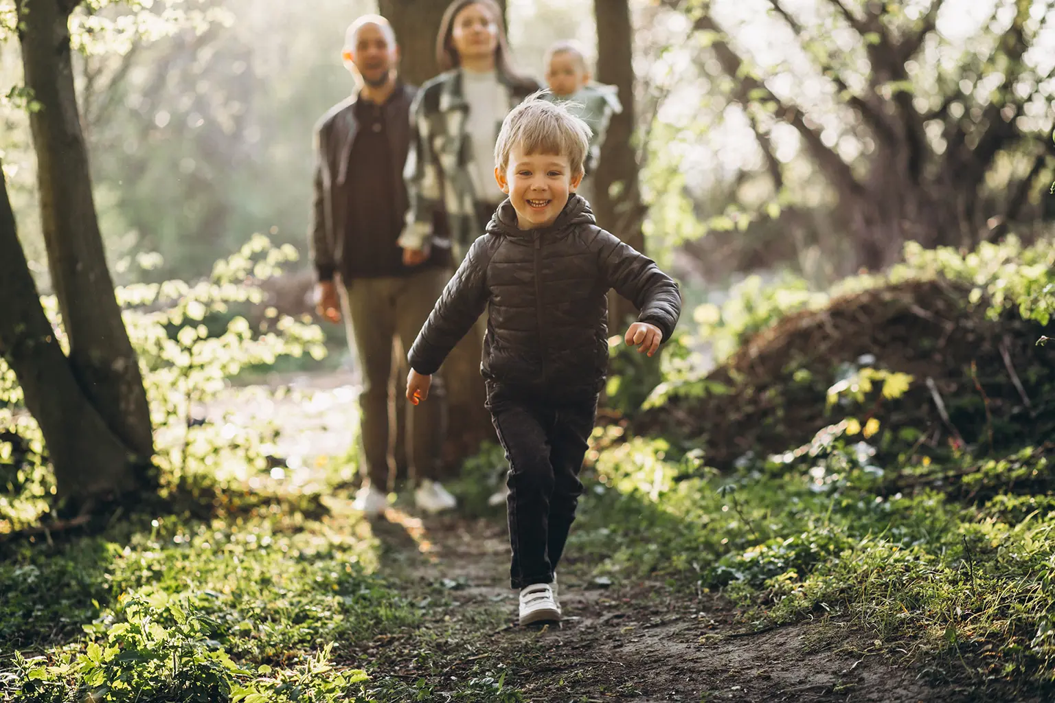 Image of a family taking a walk along a forest trail, little boy in a black jacket running ahead, smiling showcasing the general & family dentistry services offered by Parkway Family Dental.